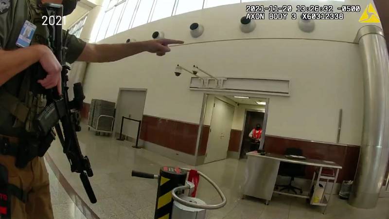 A law enforcement officer in a bullet proof vest holding an automatic weapon is seen pointing off screen in an image captured by a body camera worn by another law enforcement officer.