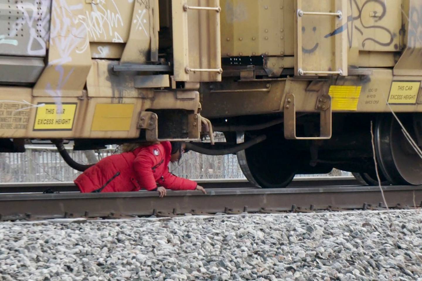 An elementary school-age child is seen crawling underneath a train car. Her hands rest on the tracks as she pulls her body across the space under the car, next to a coupling joint connecting it to the rest of the train.