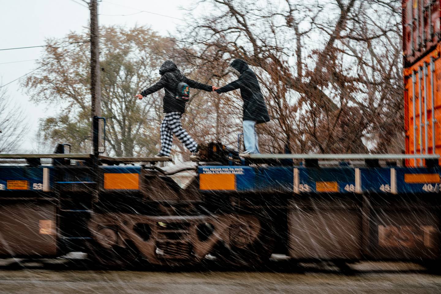 A photograph of two children holding hands as they jump over the coupling joint of a flat-bed train car. The image shows one child mid step across the gap. Shipping containers stacked two-high can be seen just next to them. They are dressed in winter clothing as light precipitation falls around them.