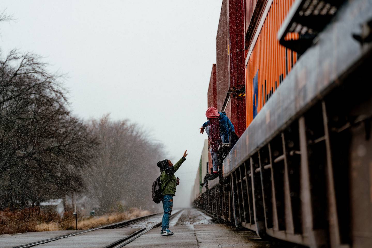 A photo shows two children, one on the ground reaching up to the other, who is in the midst of climbing down from a flatbed train car holding shipping containers stacked two-tall. The children are dressed in coats and hats as light precipitation falls around them.