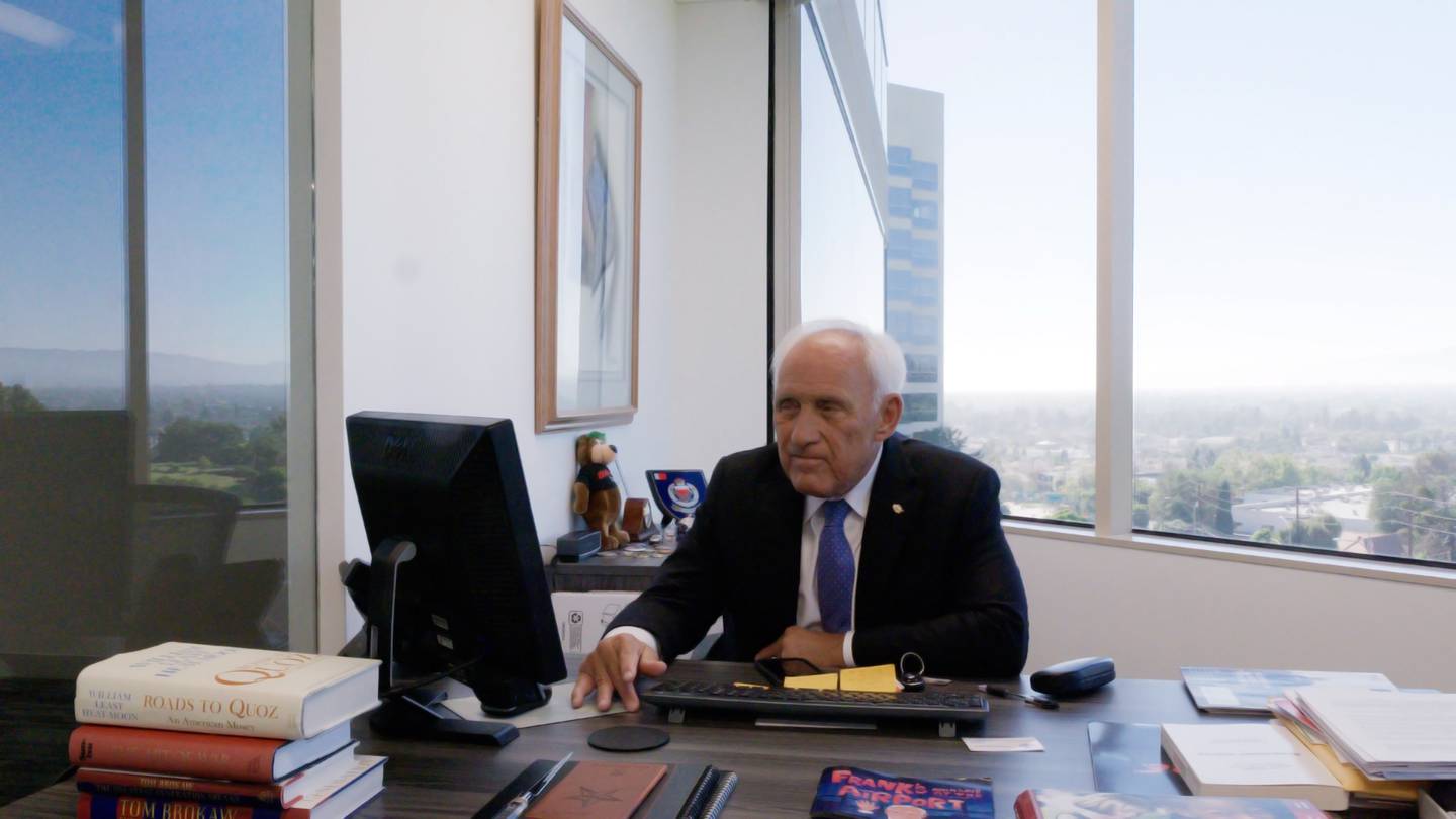 Francisco Pegueros sitting behind a desk, while looking at a computer. Behind him is a window overlooking Culver City, CA.