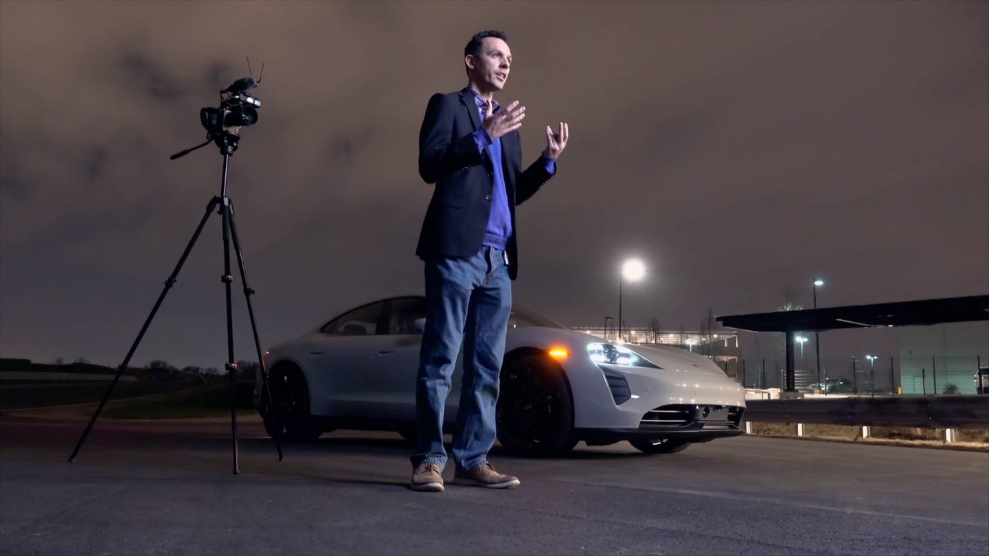 A photo of Frank Wiesmann from Porsche North America standing in front of a white Porsche with the lights on. The photo indicates Wiesmann is being interviewed, and a TV camera sits on a stand behind him.
