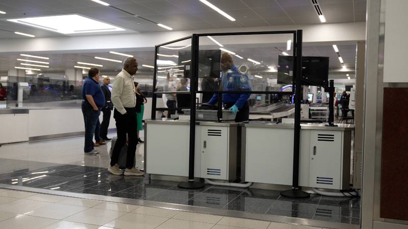 A photograph of a partial barrier at the secondary screening area of a security checkpoint. An acrylic barrier sits more than a foot above the level of a table where bags are searched, leaving a gap.